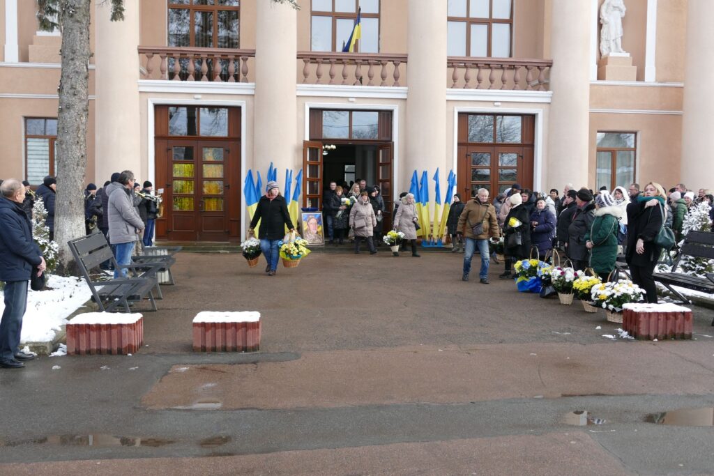 Люди з квітами під будівлею з прапорами України.