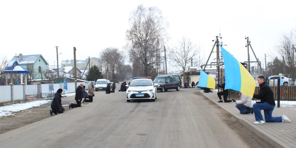 Люди на колінах тримають українські прапори вздовж дороги.