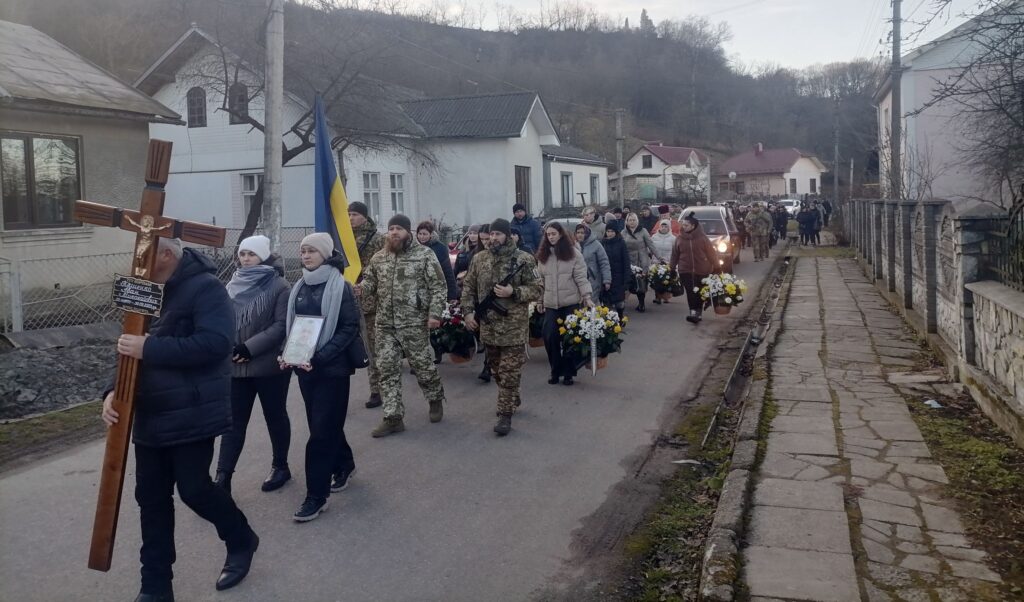 Похоронна процесія в селі з прапором і хрестом.