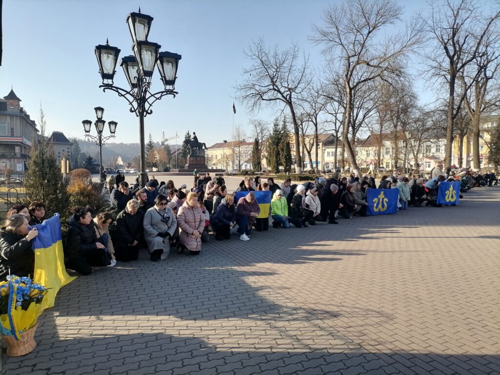 Люди на колінах на площі з українськими прапорами.