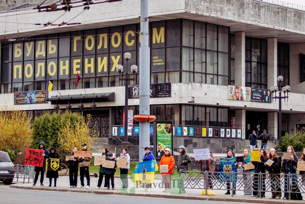 Люди тримають плакати перед будівлею з великими вікнами.