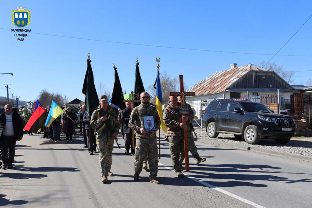 Похоронна процесія з прапорами в українському селі.
