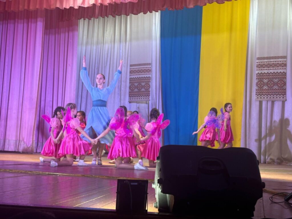 Діти танцюють на сцені з українським прапором.