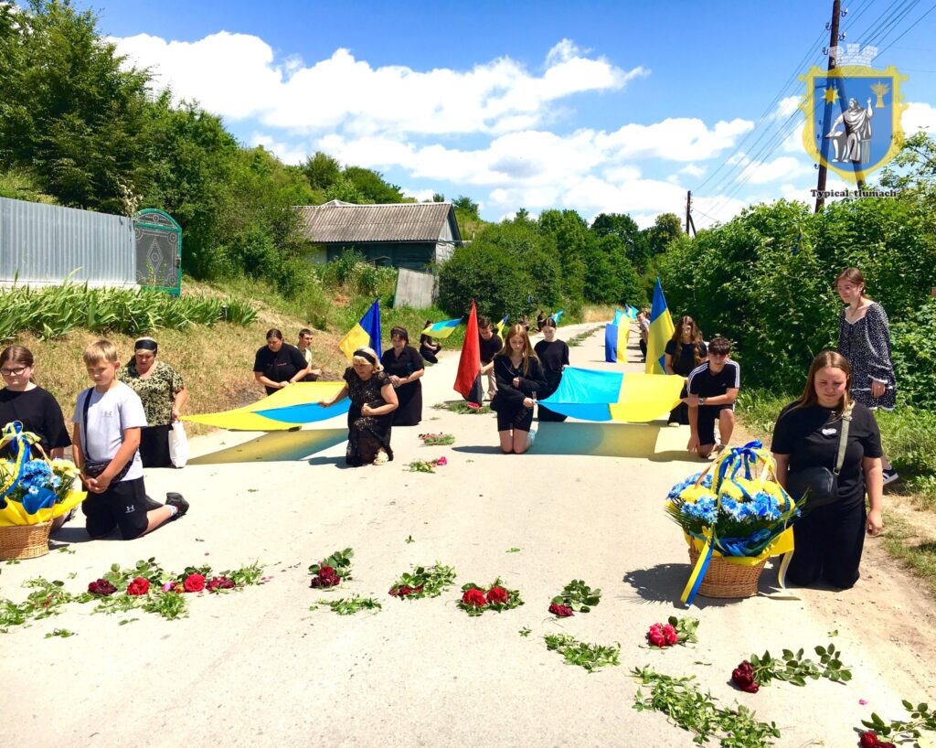 Люди на колінах з прапорами і квітами на дорозі.