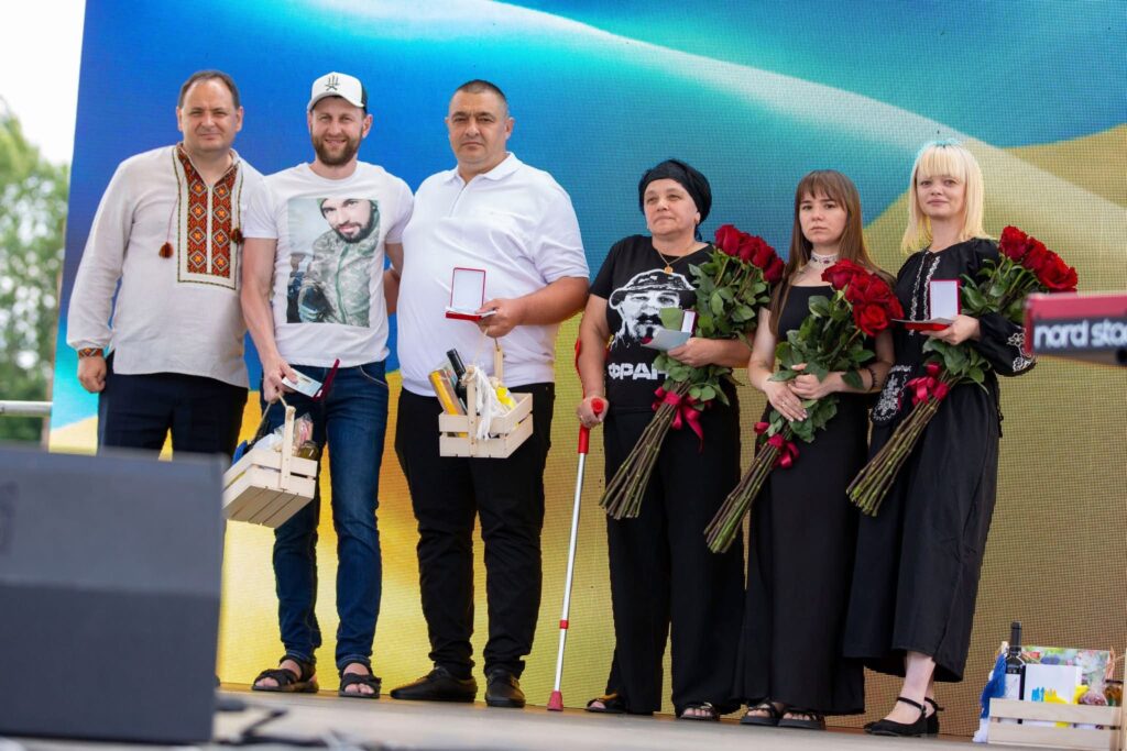 Люди з нагородами на сцені, тримають квіти.