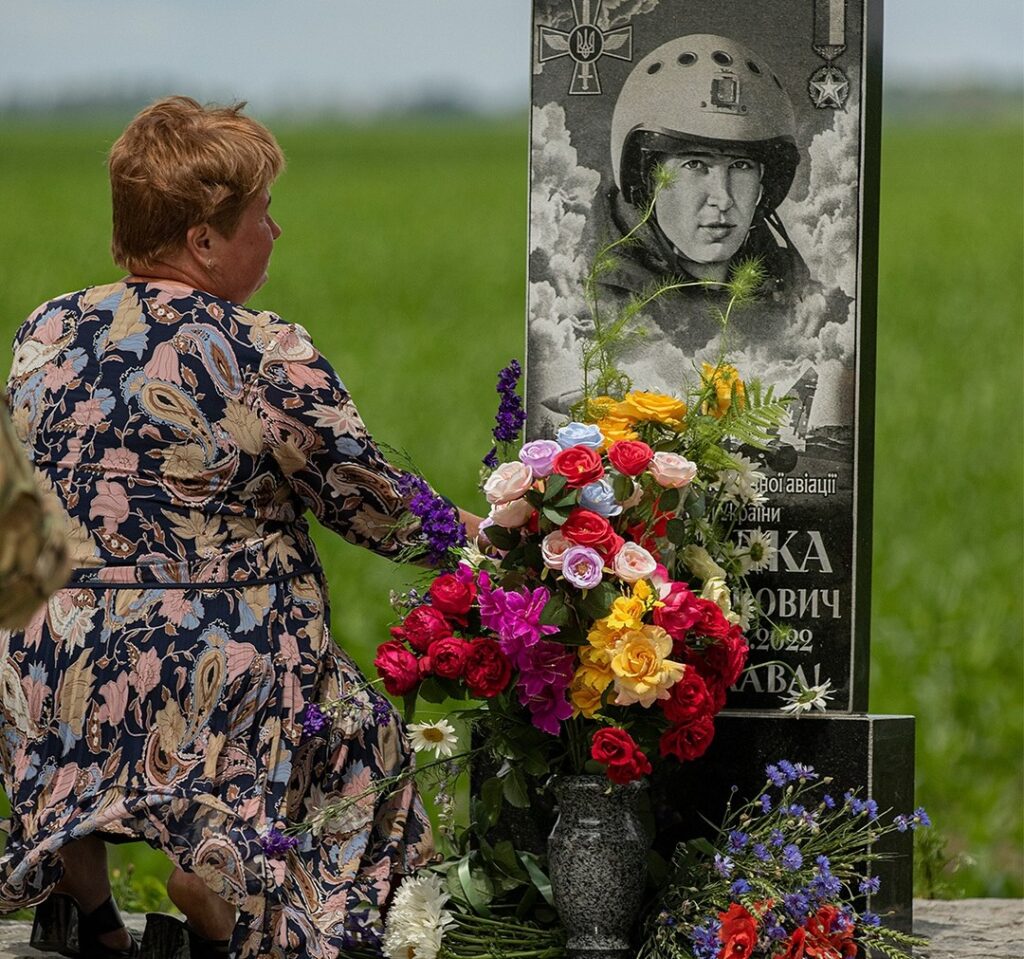 Жінка на колінах перед меморіалом з квітами.