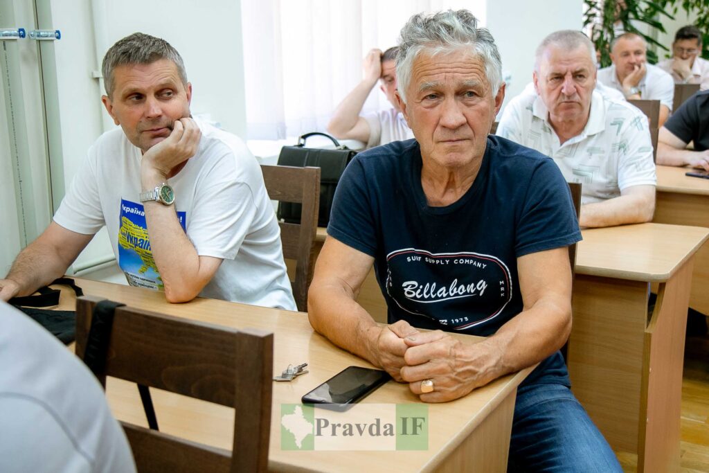 Група чоловіків сидить в аудиторії за столами.