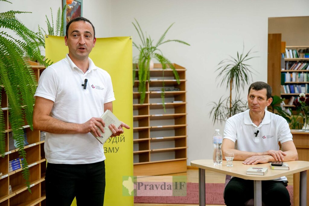 Два чоловіки в бібліотеці, один стоїть, один сидить.