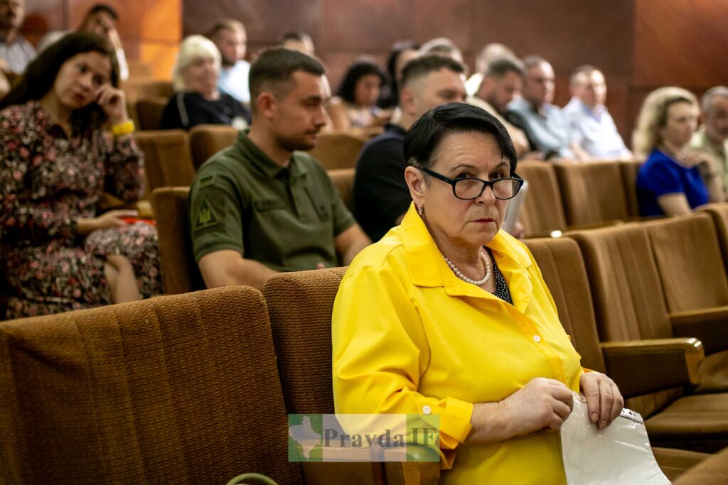 Жінка в жовтому на конференції, люди на фоні.