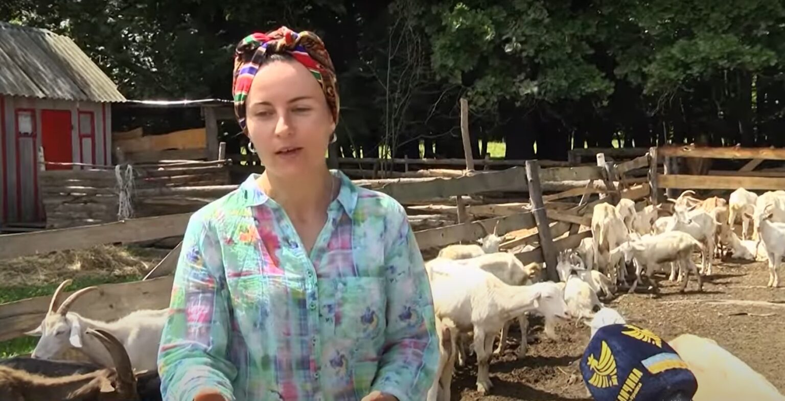 Жінка на фермі з козами на задньому плані.