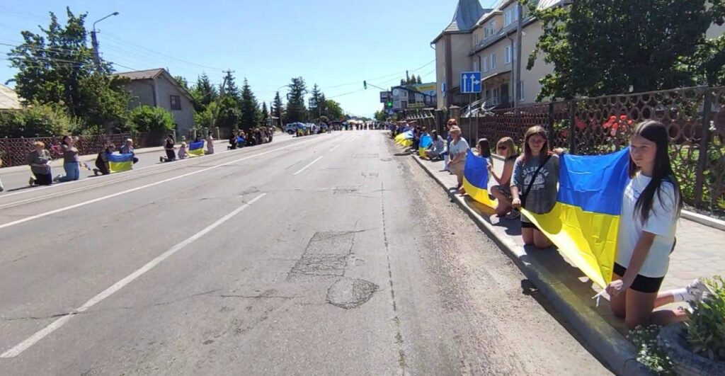 Люди з прапорами України стоять вздовж дороги.