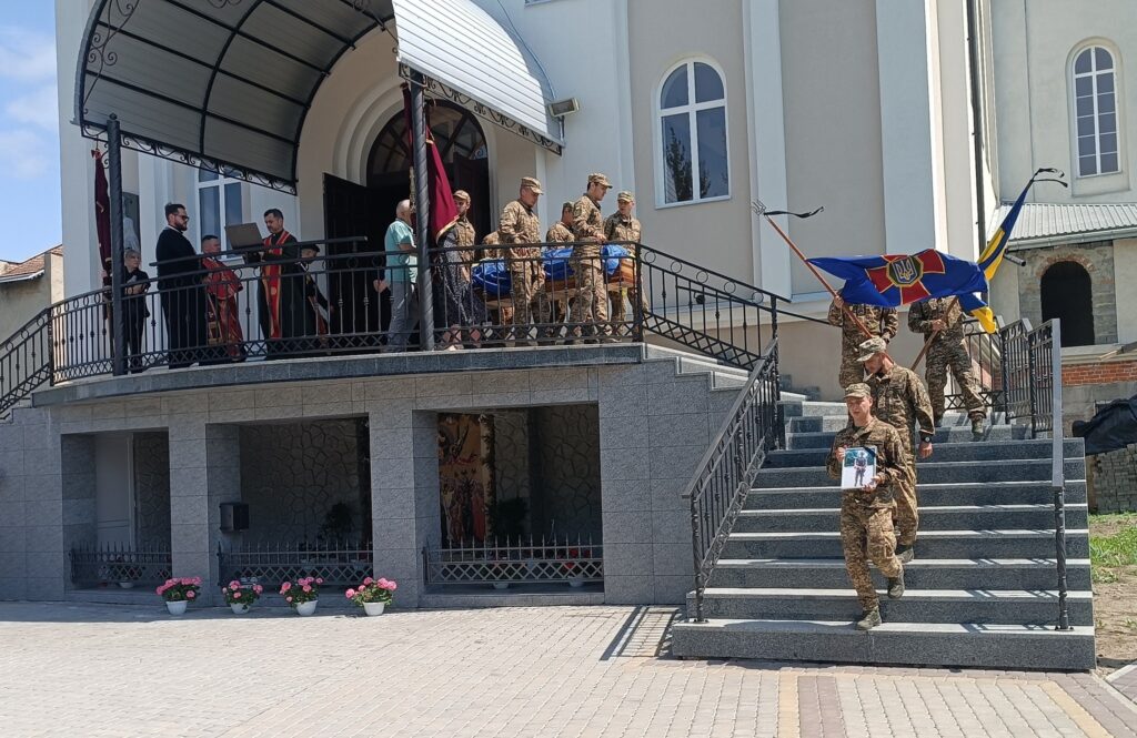 Військові несуть труну під прапором біля церкви