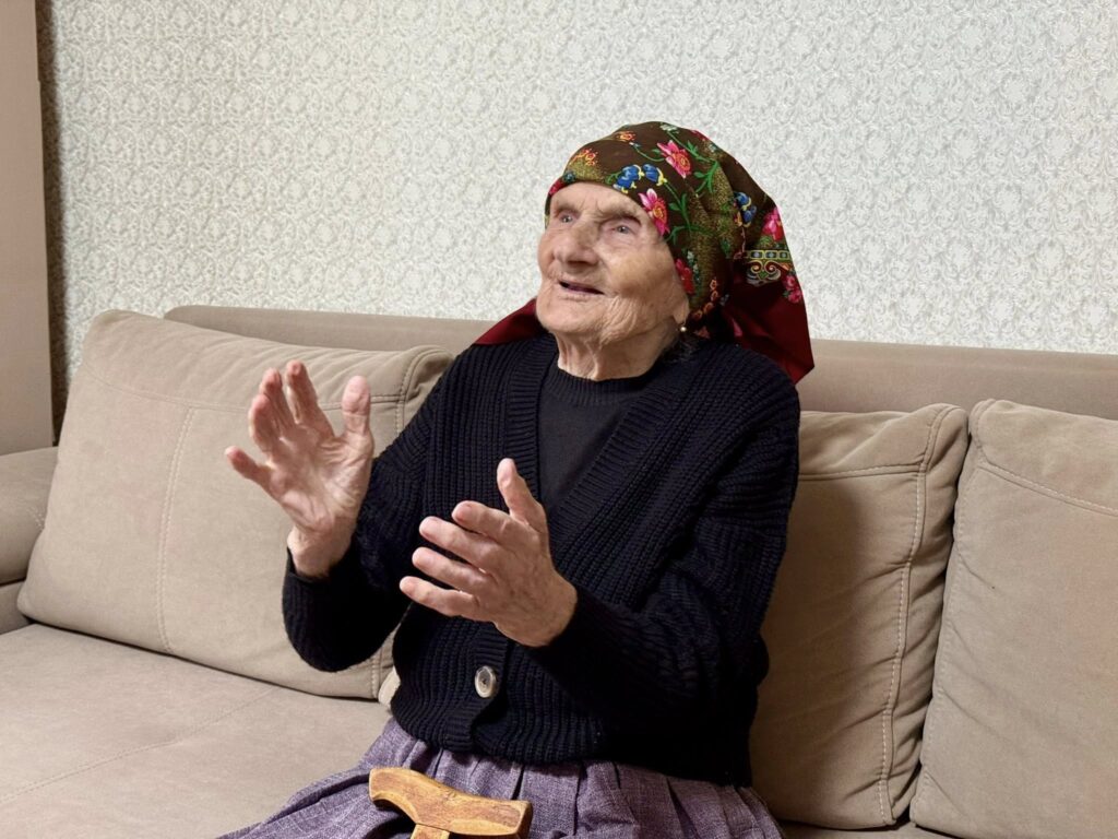 Старша жінка в хустці сидить на дивані.