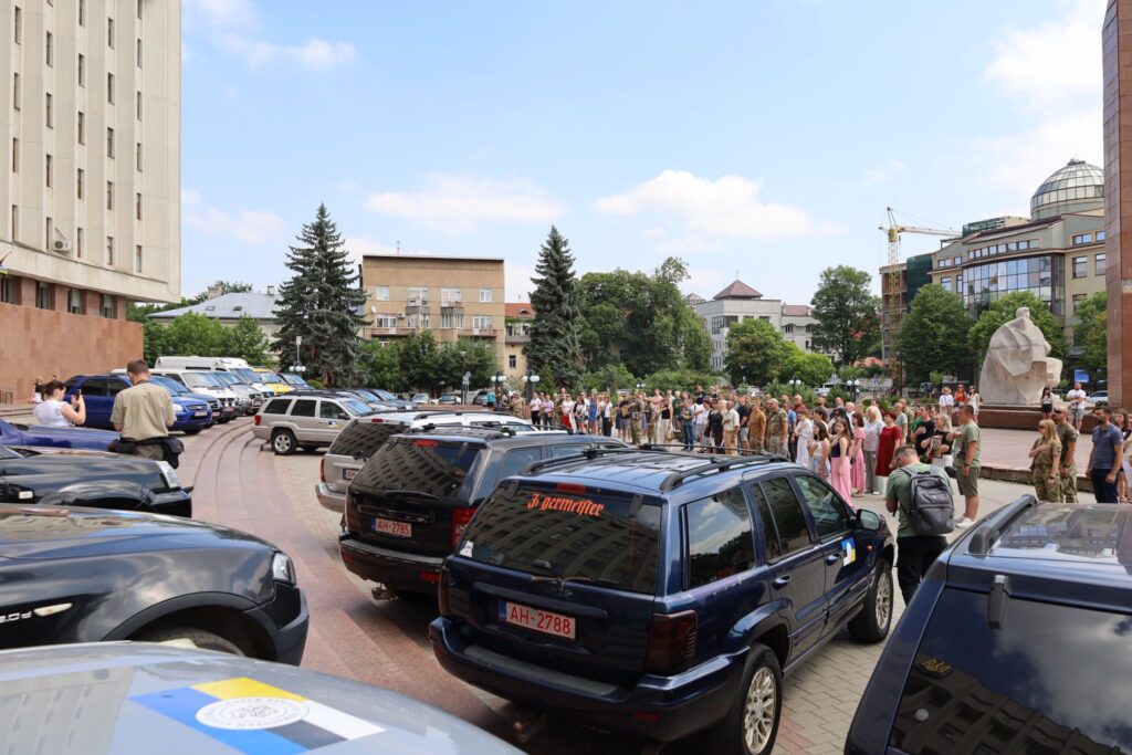 Збір людей біля будівлі з припаркованими автомобілями.
