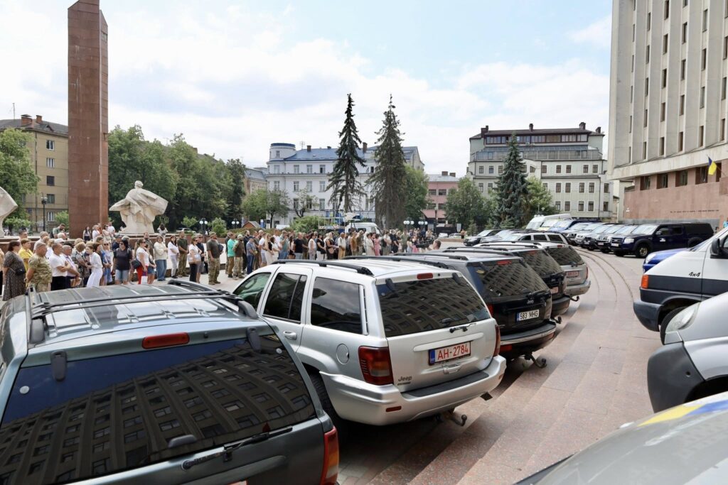 Люди в черзі на площі з автівками.