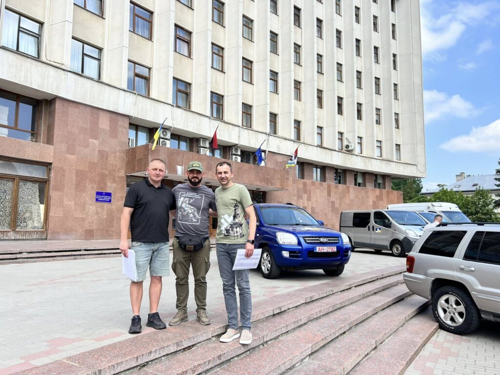 Троє людей перед адміністративною будівлею з автомобілями.