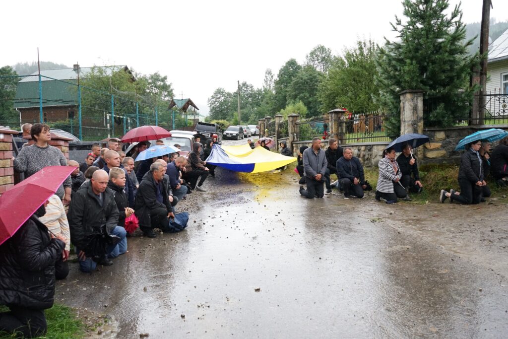 Люди з прапором України стоять на колінах під дощем.
