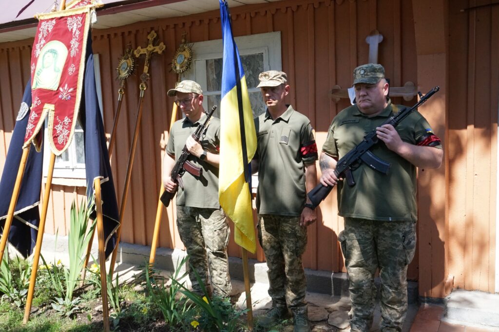 Українські солдати з прапором біля дерев'яної будівлі.