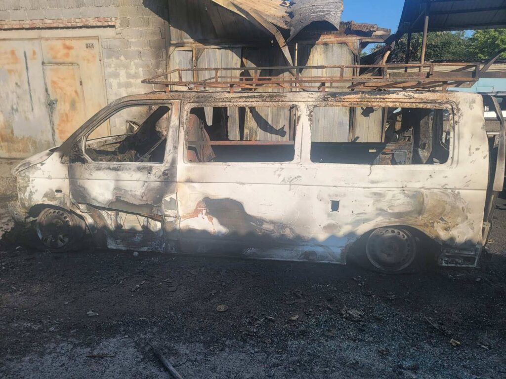 Згорілий фургон на фоні гаража та дерева.