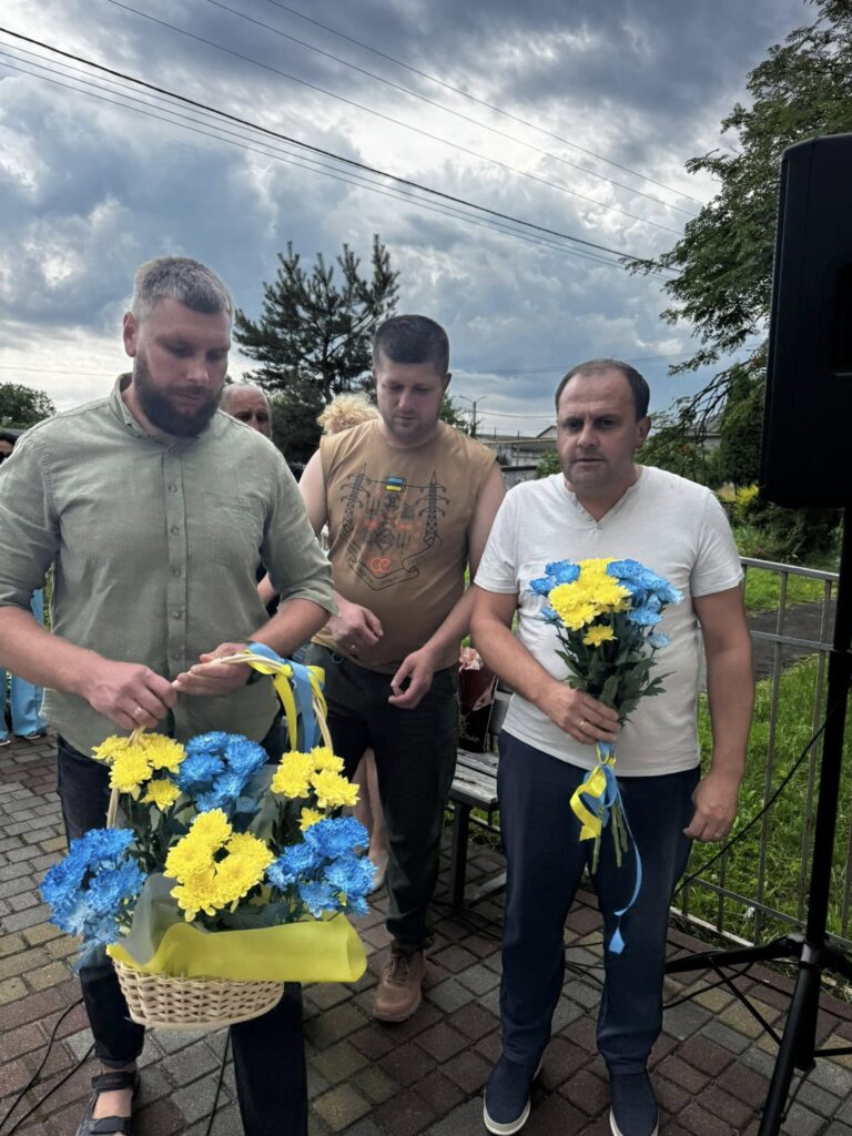 Три чоловіка з жовто-блакитними квітами під хмарами.
