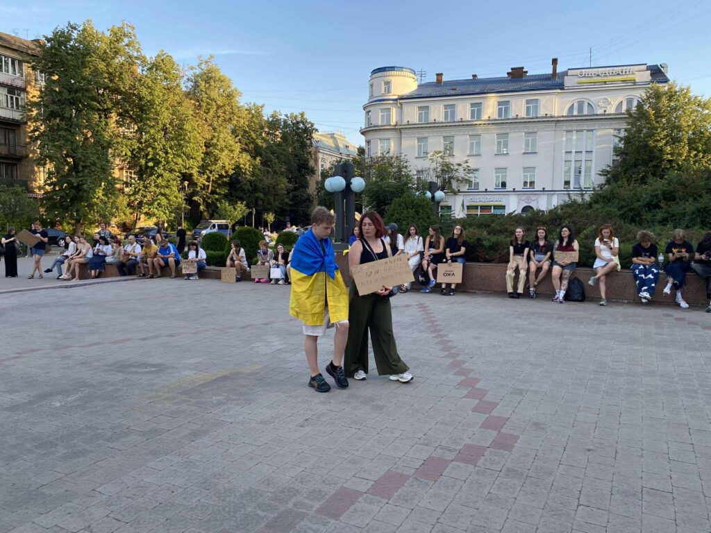Люди на площі з плакатами, один загорнутий у прапор.