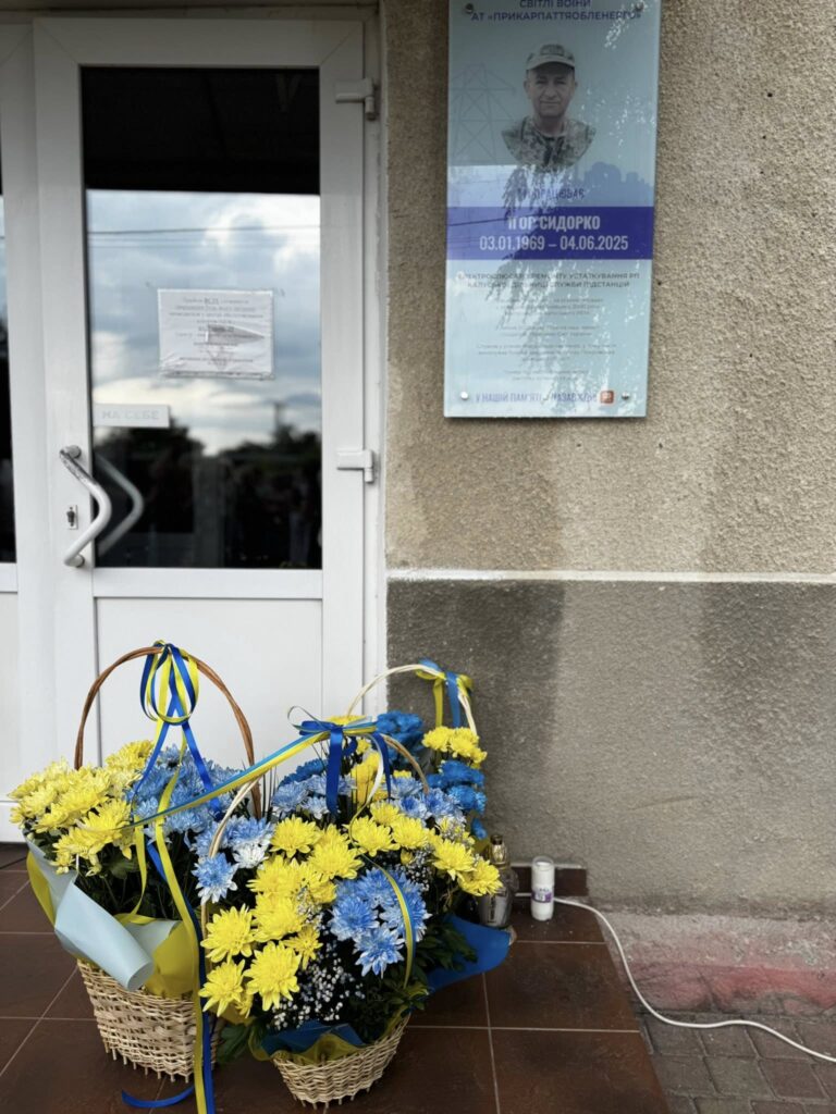 Пам'ятна дошка та жовто-блакитні квіти біля входу.