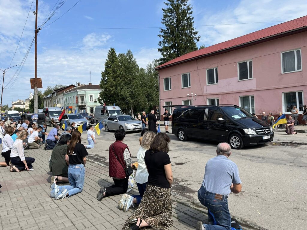 Люди на колінах шанують похоронний кортеж, прапори навколо.