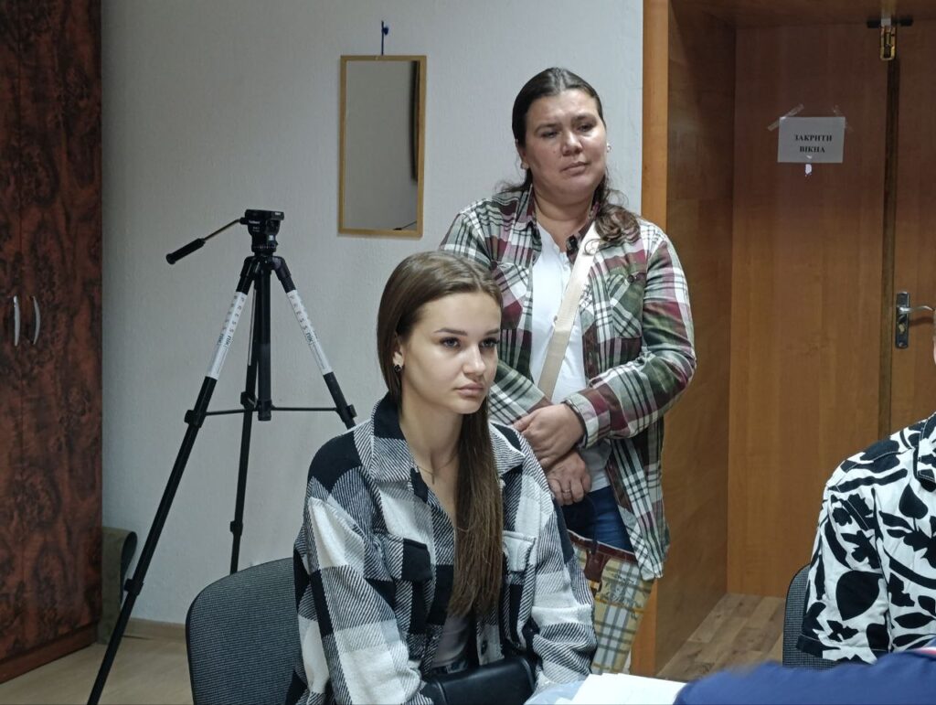 Дві жінки сидять і стоять у кімнаті.