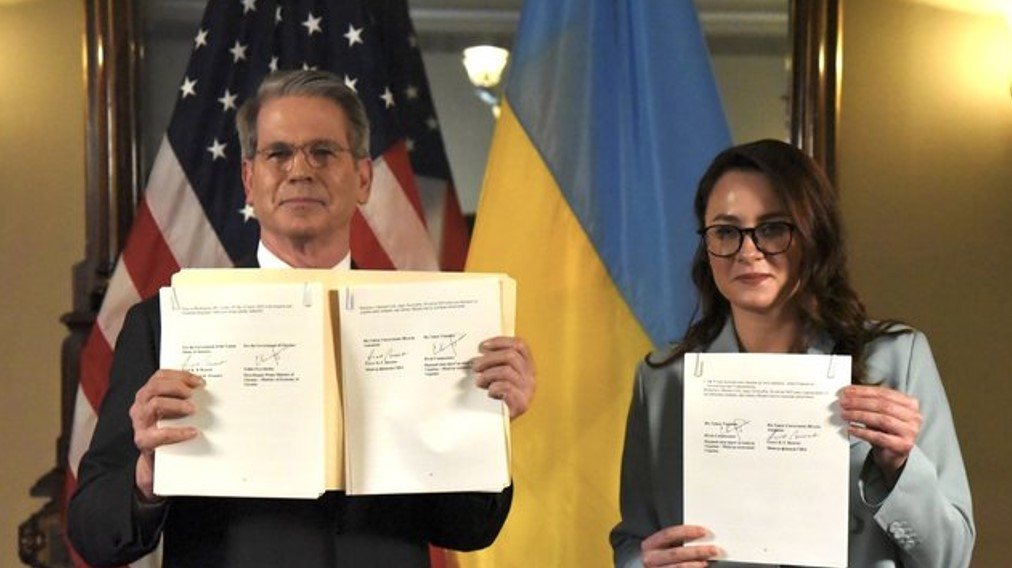 Люди тримають підписані документи перед прапорами США та України.