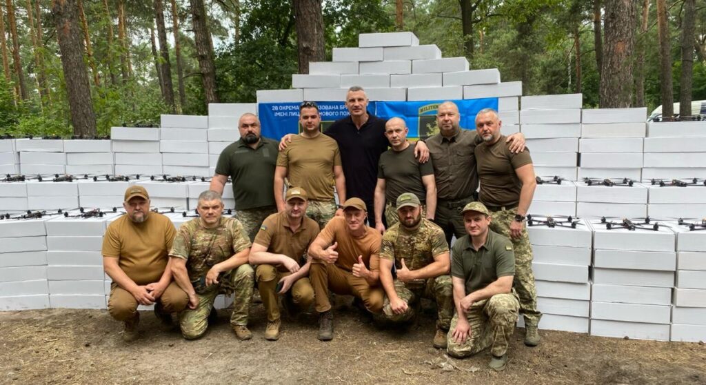 Група чоловіків перед білими коробками в лісі.