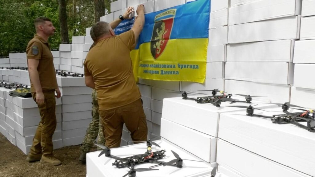 Військові упаковують коробки з дронами біля прапора.