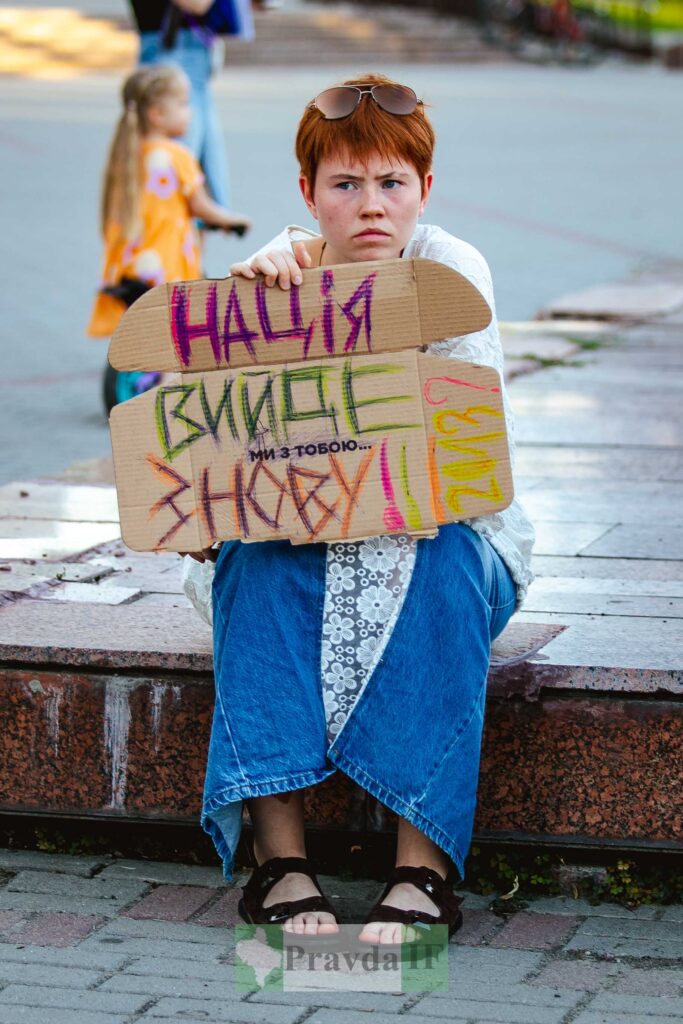 Жінка сидить з картонним плакатом на вулиці.