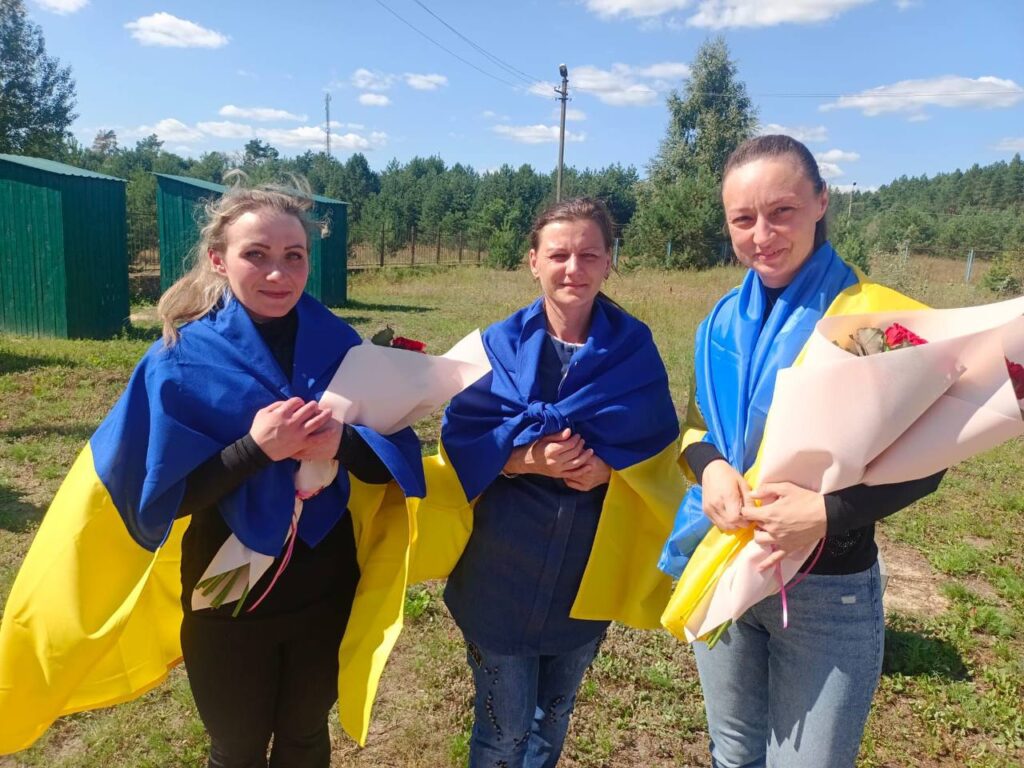 Жінки з квітами, загорнуті в українські прапори.