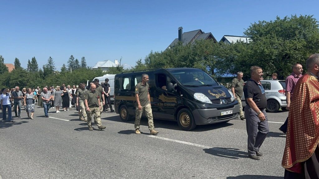 Похоронна процесія вулицею за чорним мікроавтобусом.