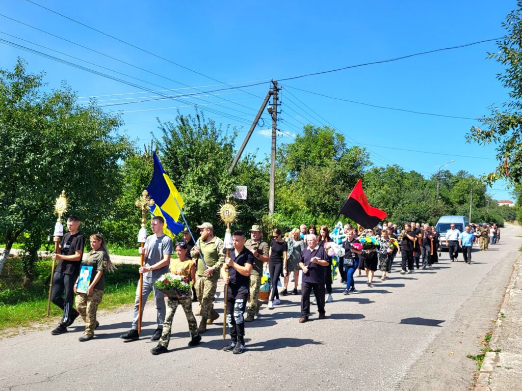 Люди йдуть вулицею з прапорами та квітами.