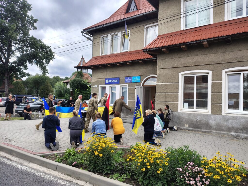 Люди з прапорами України на колінах біля будівлі.