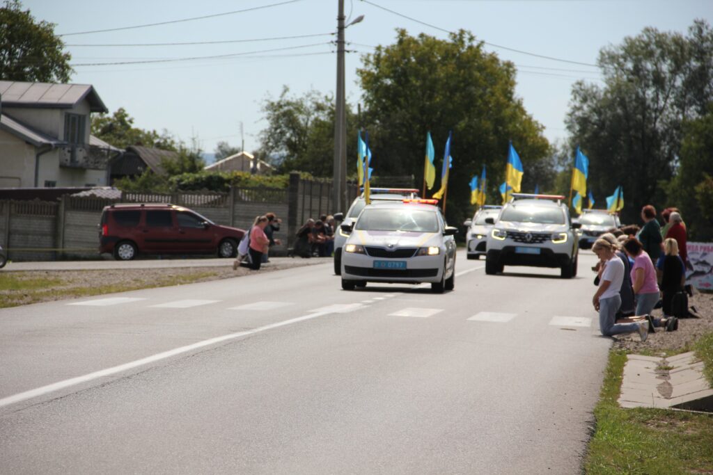 Автомобілі з прапорами України на дорозі, люди на колінах.