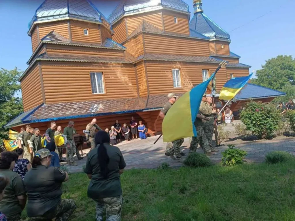 Церемонія біля дерев'яної церкви, люди з прапорами України.