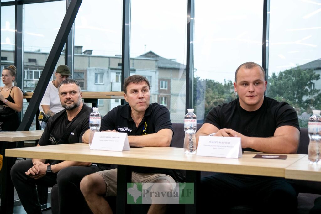 Пресконференція чоловіків у залі зі скляними вікнами.
