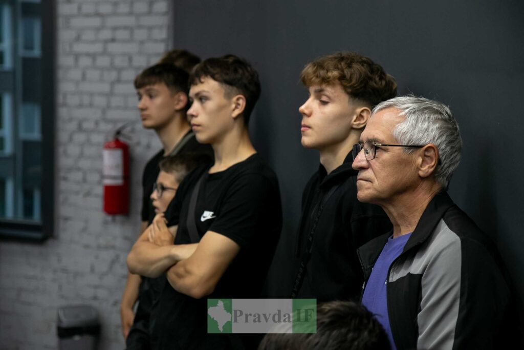Група людей, стоять у приміщенні, уважно слухають.