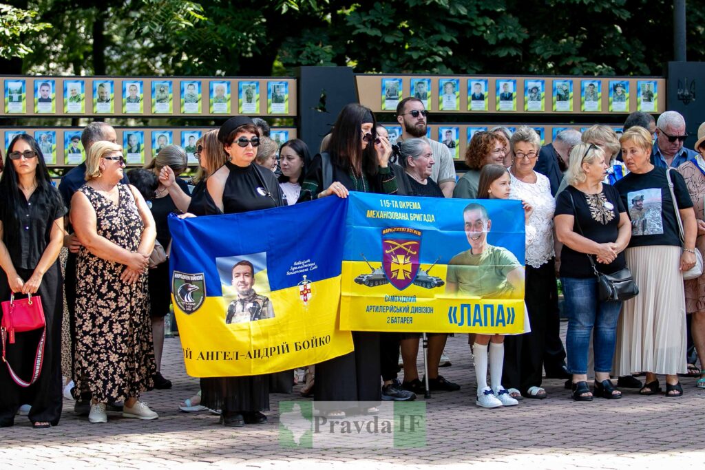 Люди на меморіалі з прапорами та фото героїв.