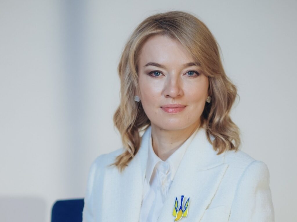 Жінка у білому піджаку, світле волосся.