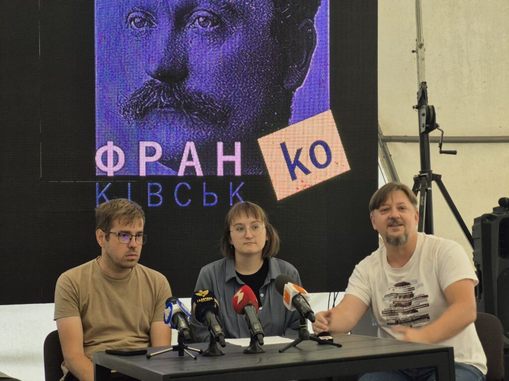 Троє спікерів на пресконференції, портрет Франка на екрані.