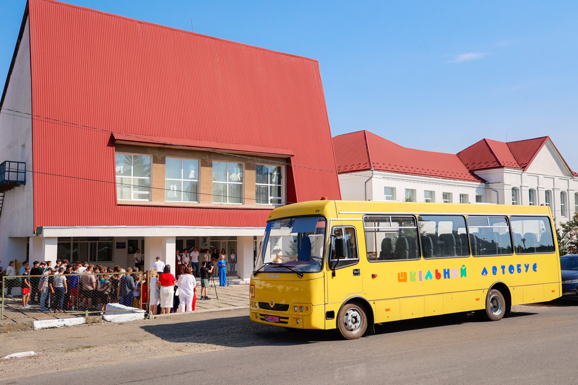 Жовтий шкільний автобус біля школи з червоним дахом.