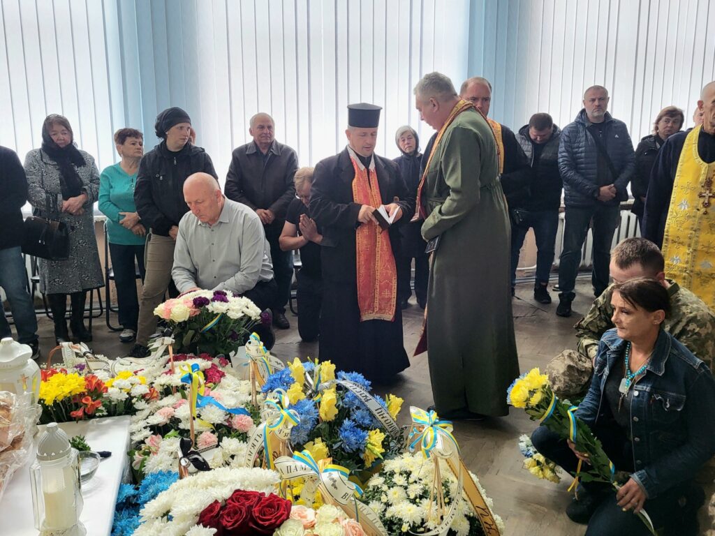Люди на похороні з квітами й священником.
