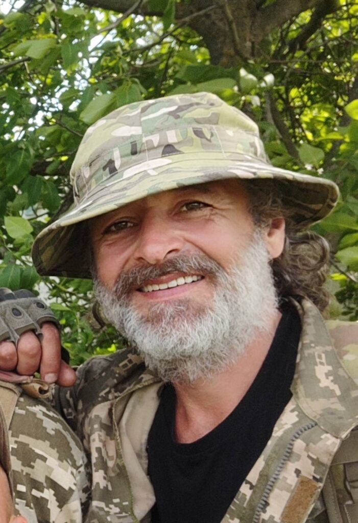 Усміхнений чоловік у камуфляжі та капелюсі на природі.