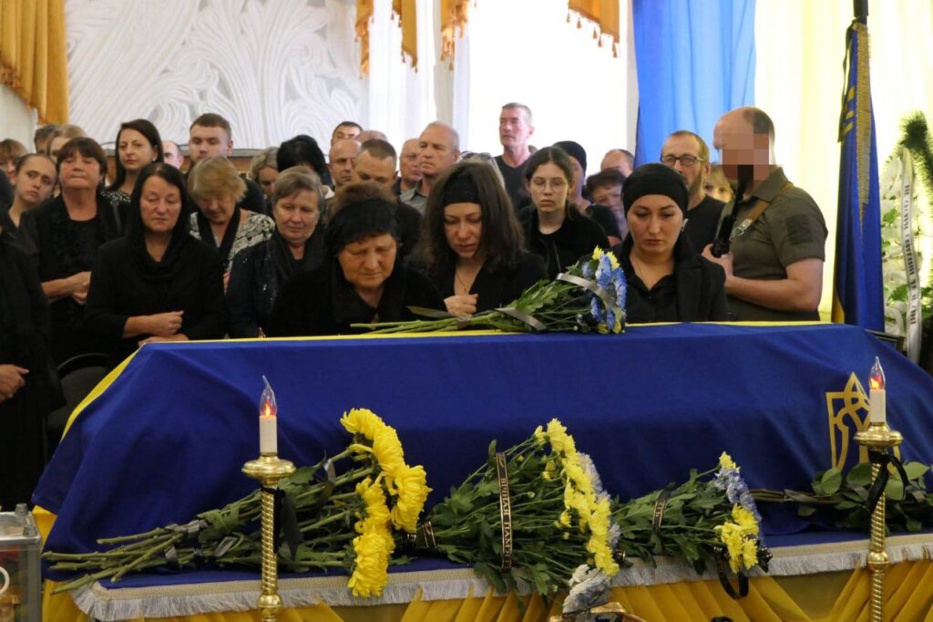 Люди у траурному одязі біля труни з квітами.