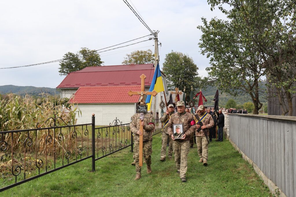 Військовий похорон з українським прапором, пейзаж села.