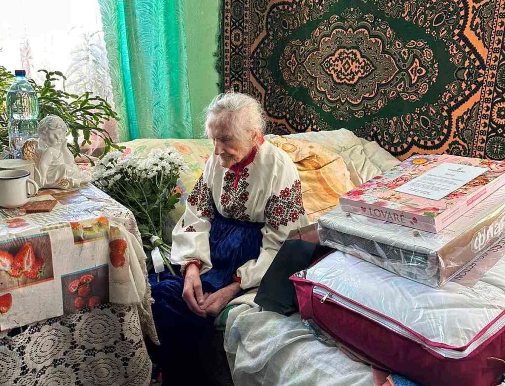 Старша жінка в кімнаті з подарунками та квітами.