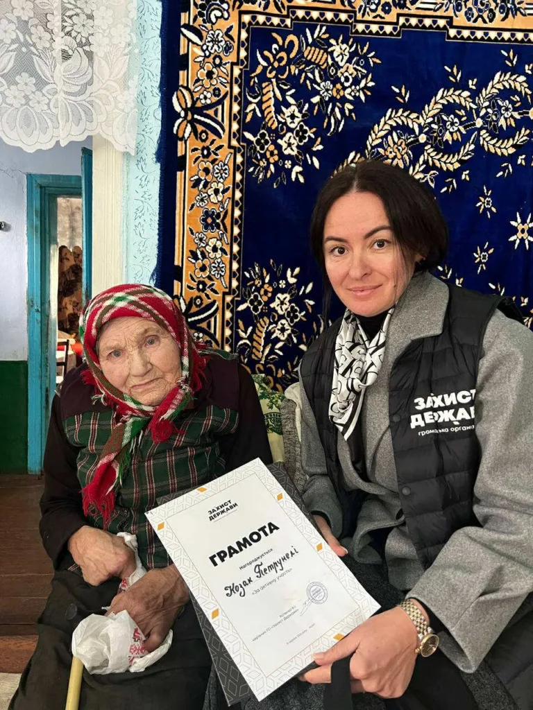 Жінка вручає грамоту літній жінці в хустці